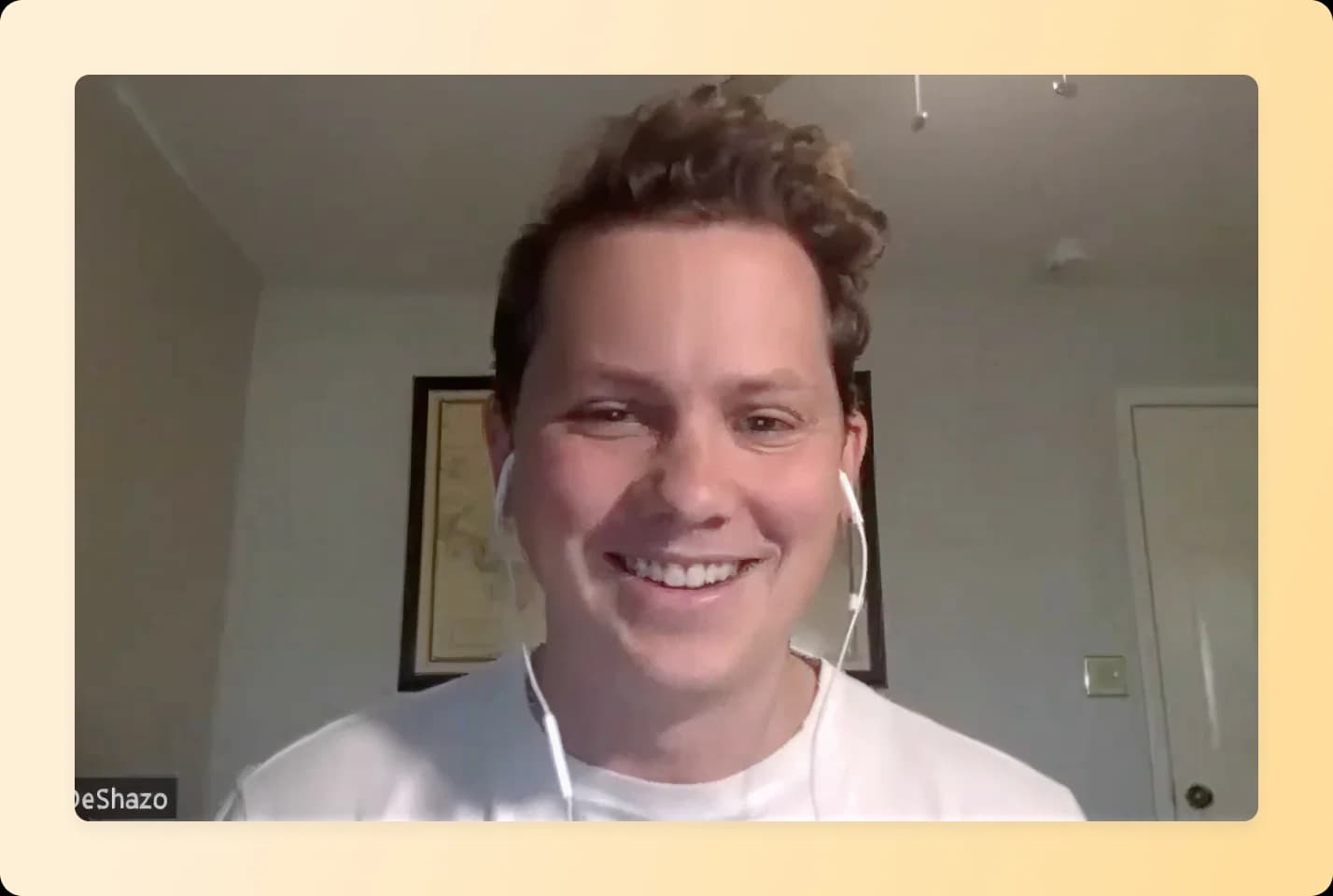 Smiling person with curly hair wearing white t-shirt and earbuds during video call in home office with framed artwork